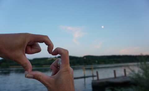 Hände formen ein Herz vor einem ruhigen See bei Abenddämmerung