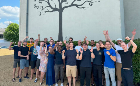 Gruppenfoto des carmasec-Teams in Freizeitkleidung, jubelnd und mit erhobenen Armen vor einem Baumwandgemälde – carmasec Week 2024